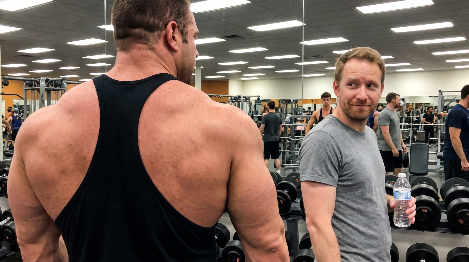 A man in a grey t-shirt holding a water bottle glances, amused, at the huge traps and shoulders of a bodybuilder in a black stringer tank top in a busy gym