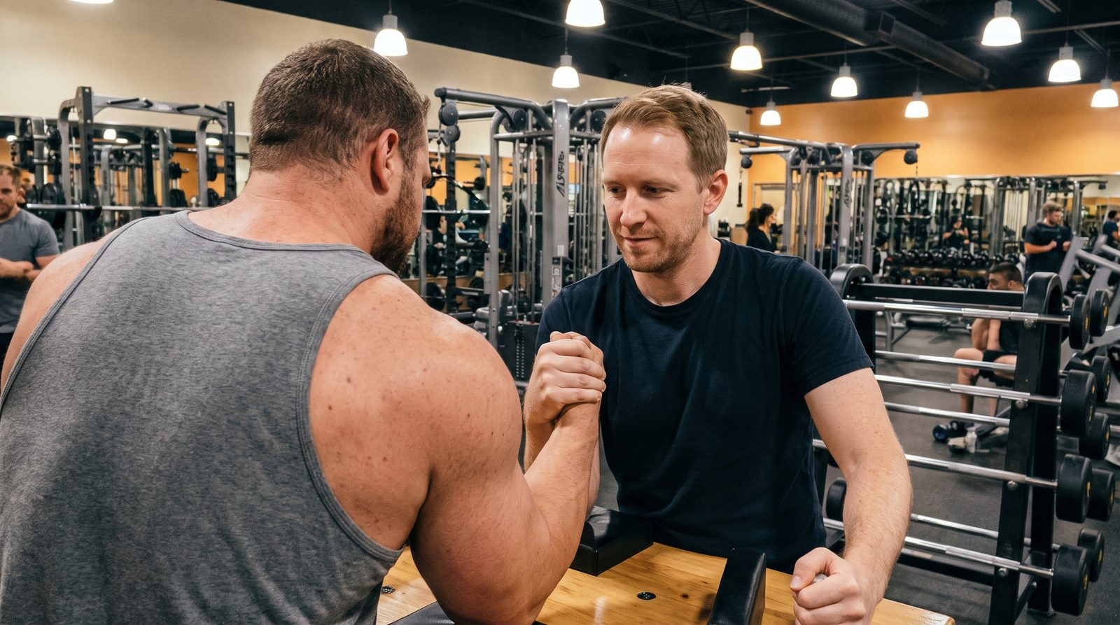 A lean man in a navy t-shirt arm wrestling a much bigger, heavily muscled man in a grey tank top at a table in a busy gym