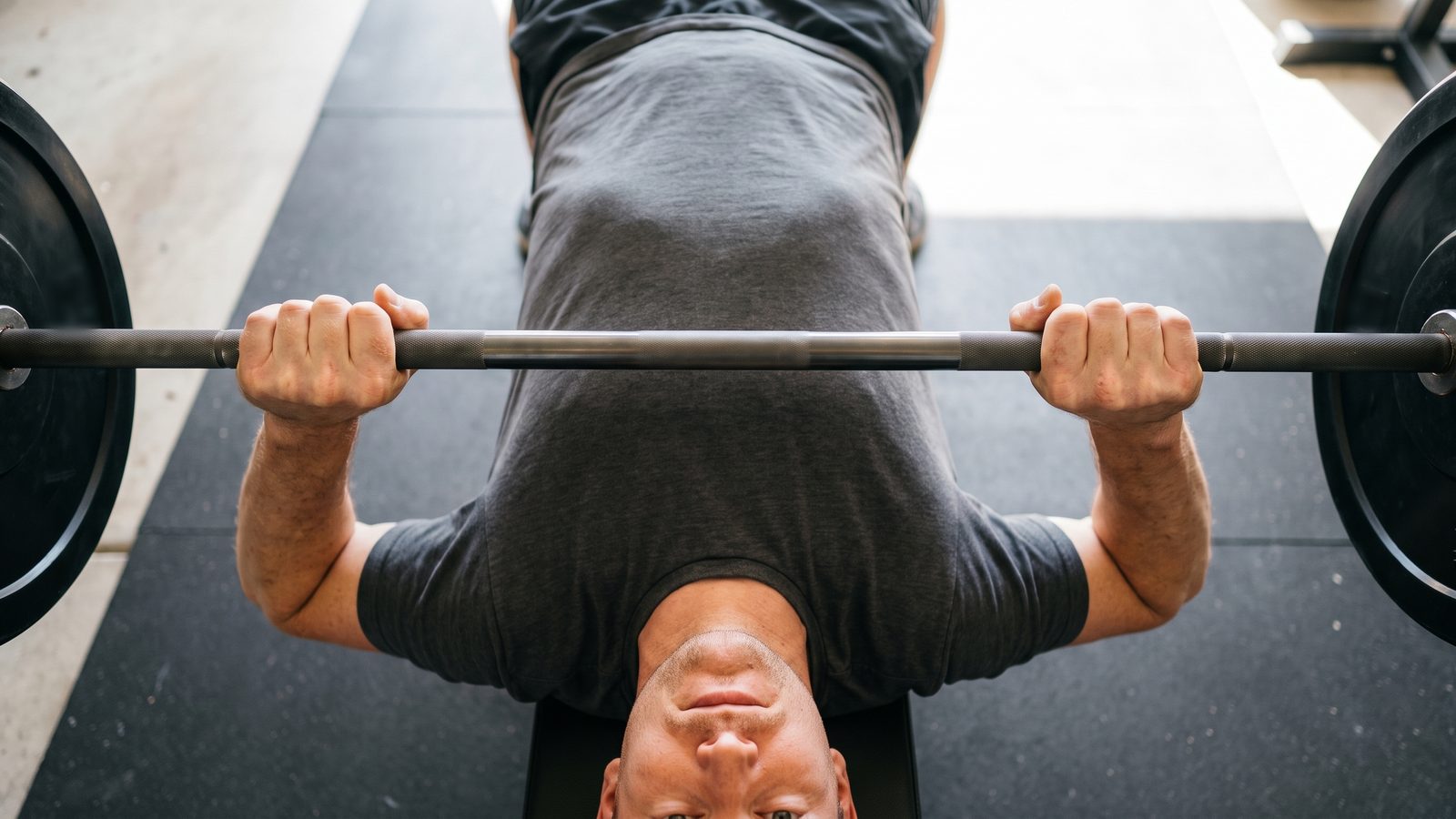 Looking down the length of a flat bench at a man lying on it, both hands gripping a barbell wide over his chest