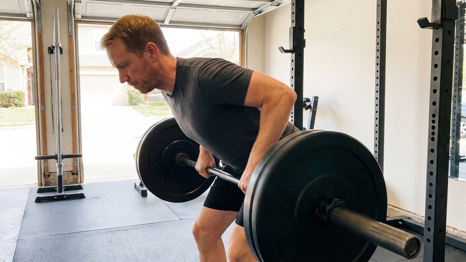 A man doing a bent-over barbell row in a garage gym, torso leaning forward with a flat back, pulling the bar to his lower ribs, seen from the side