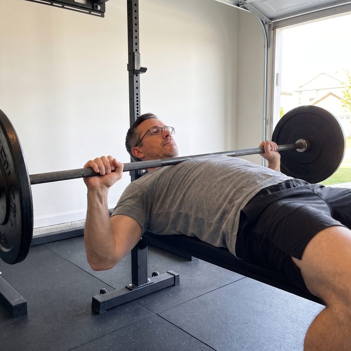 A man in glasses at the bottom of a bench press in a garage gym, a straight bar with one plate a side resting on his chest, wide grip
