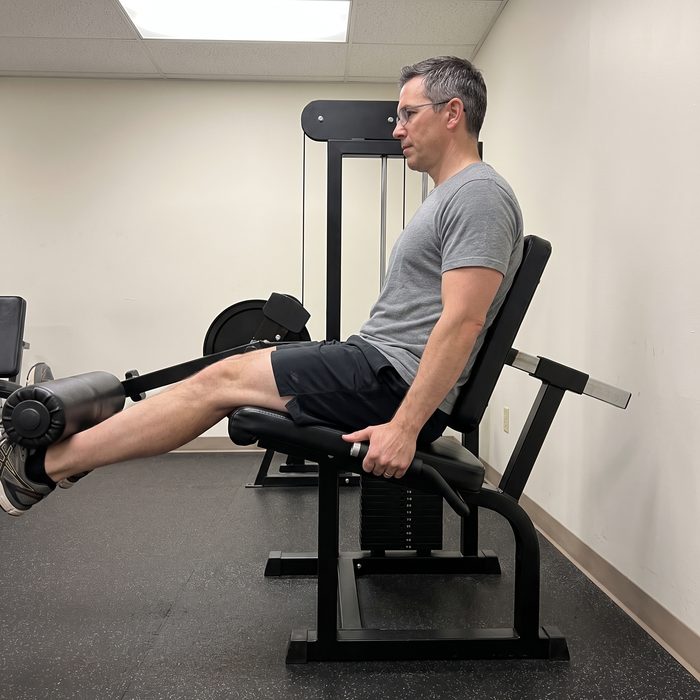 A man in glasses seated on a leg curl machine with his hips bent and legs out in front, the hamstrings at their longest