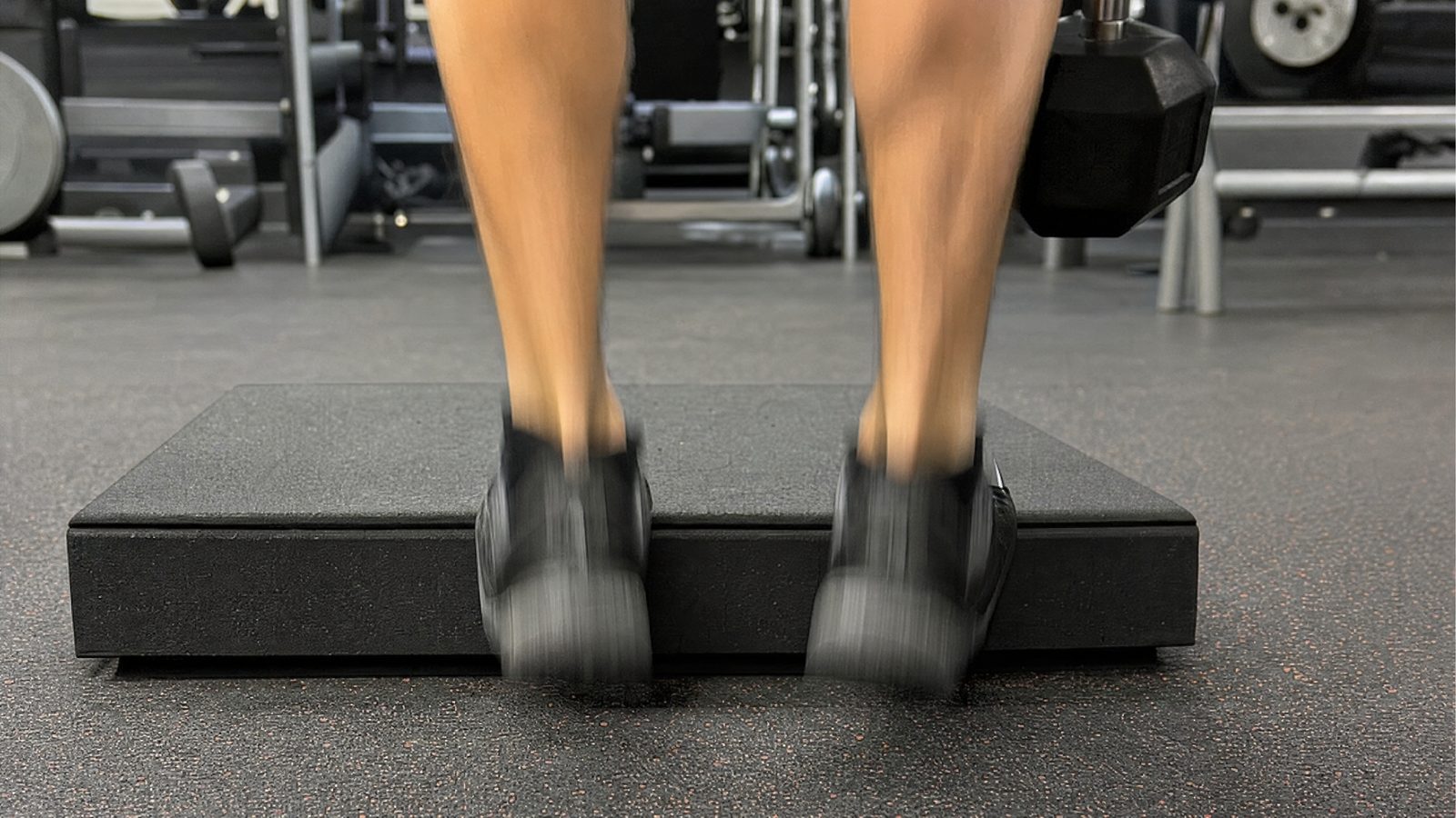 Close-up from behind of two feet in black training shoes on the edge of a black rubber block in a gym, the heels and calves blurred with motion as they bounce through a calf raise, a hex dumbbell hanging beside them