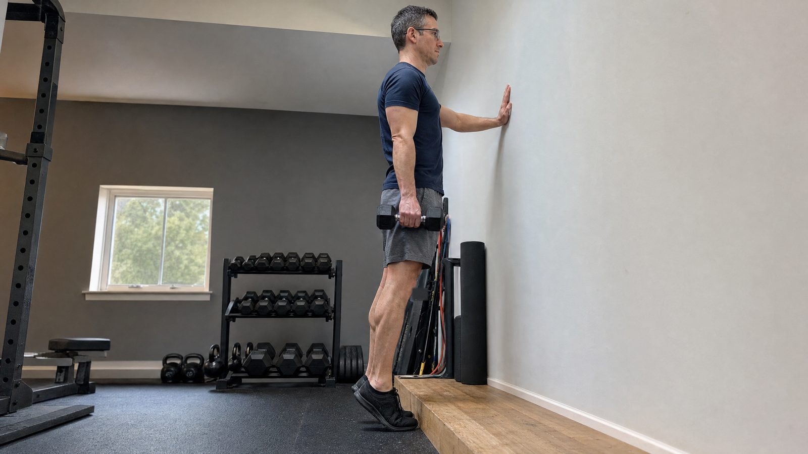 A man in glasses and a navy t-shirt standing on the edge of a low wooden step in a home gym, one hand on the wall, a hex dumbbell in the other, heels dropped below the step at the bottom of a calf raise