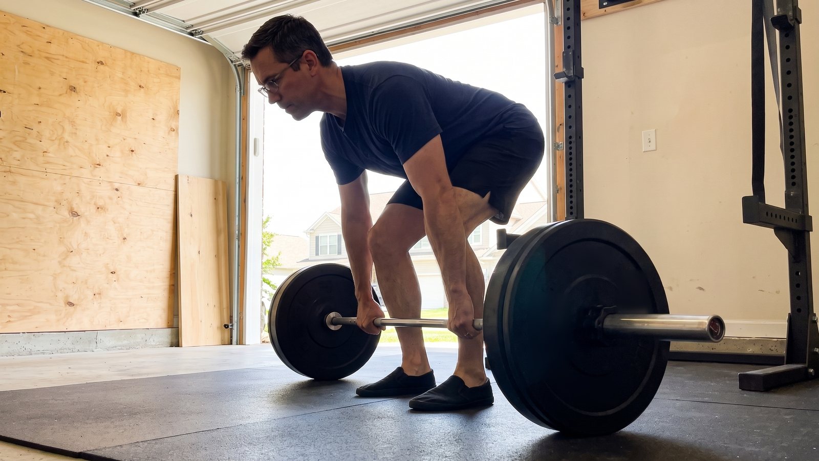 A man in glasses at the start of a conventional barbell deadlift in a garage gym, seen from the side, arms straight and hips set a little above his knees