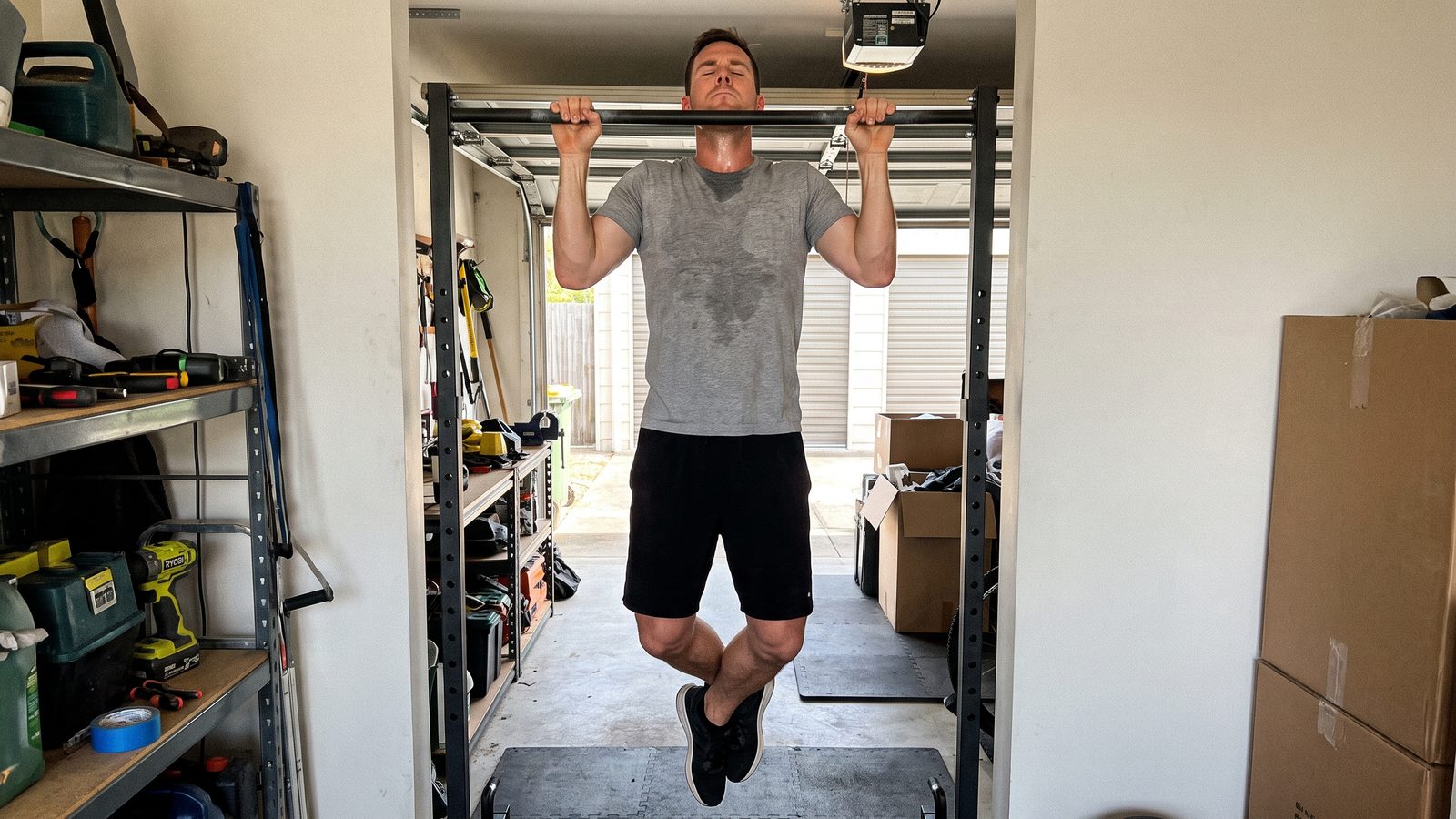 A man hanging at the top of a pull-up on a bar in a garage, chin level with the bar and elbows starting to open