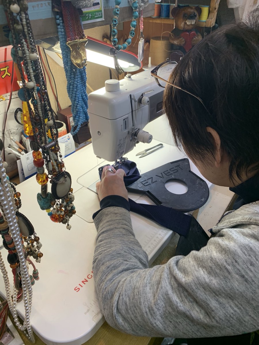 A tailor sewing navy webbing strapping on a domestic sewing machine, with a black 3D printed vest plate on the table beside the machine