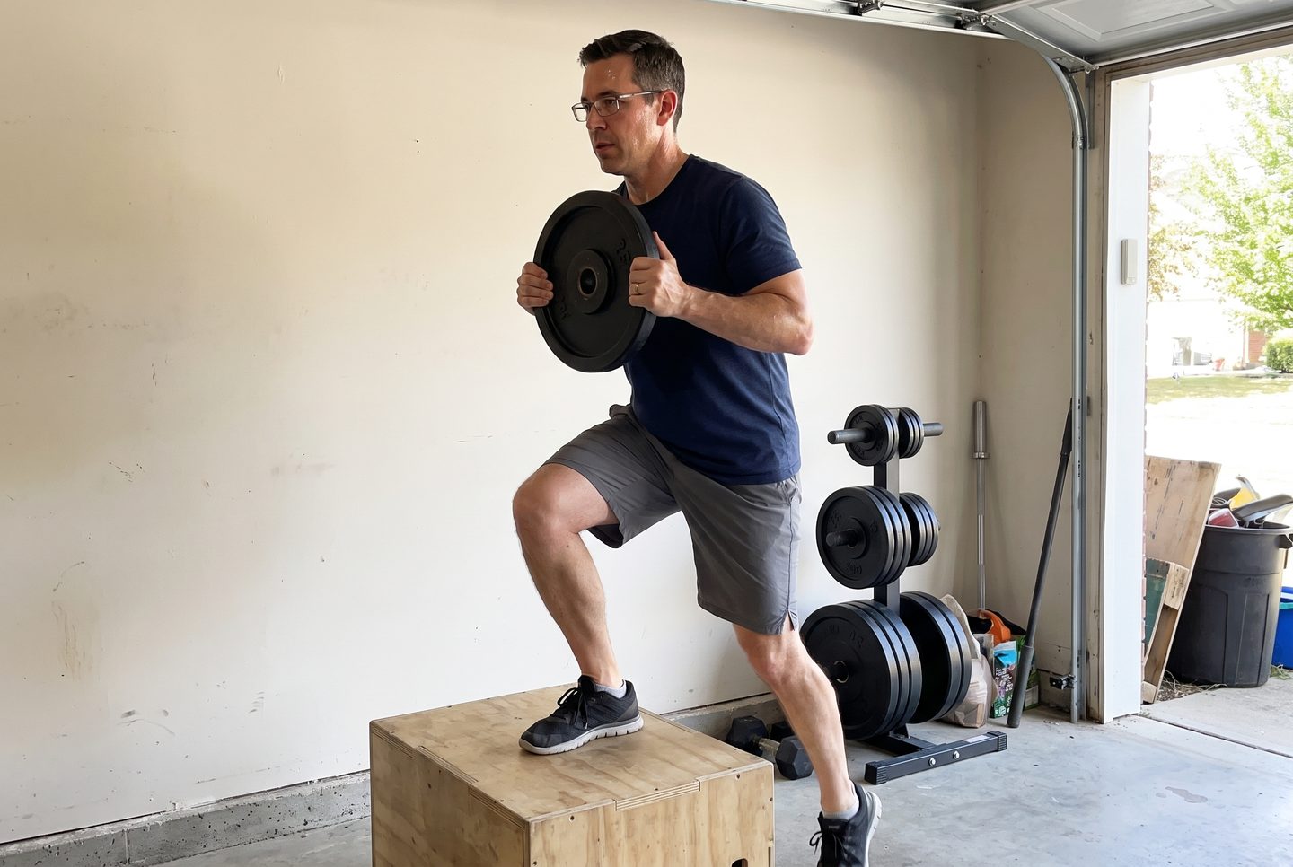 A man in his forties stepping up onto a wooden box holding a single iron plate against his chest in a garage gym