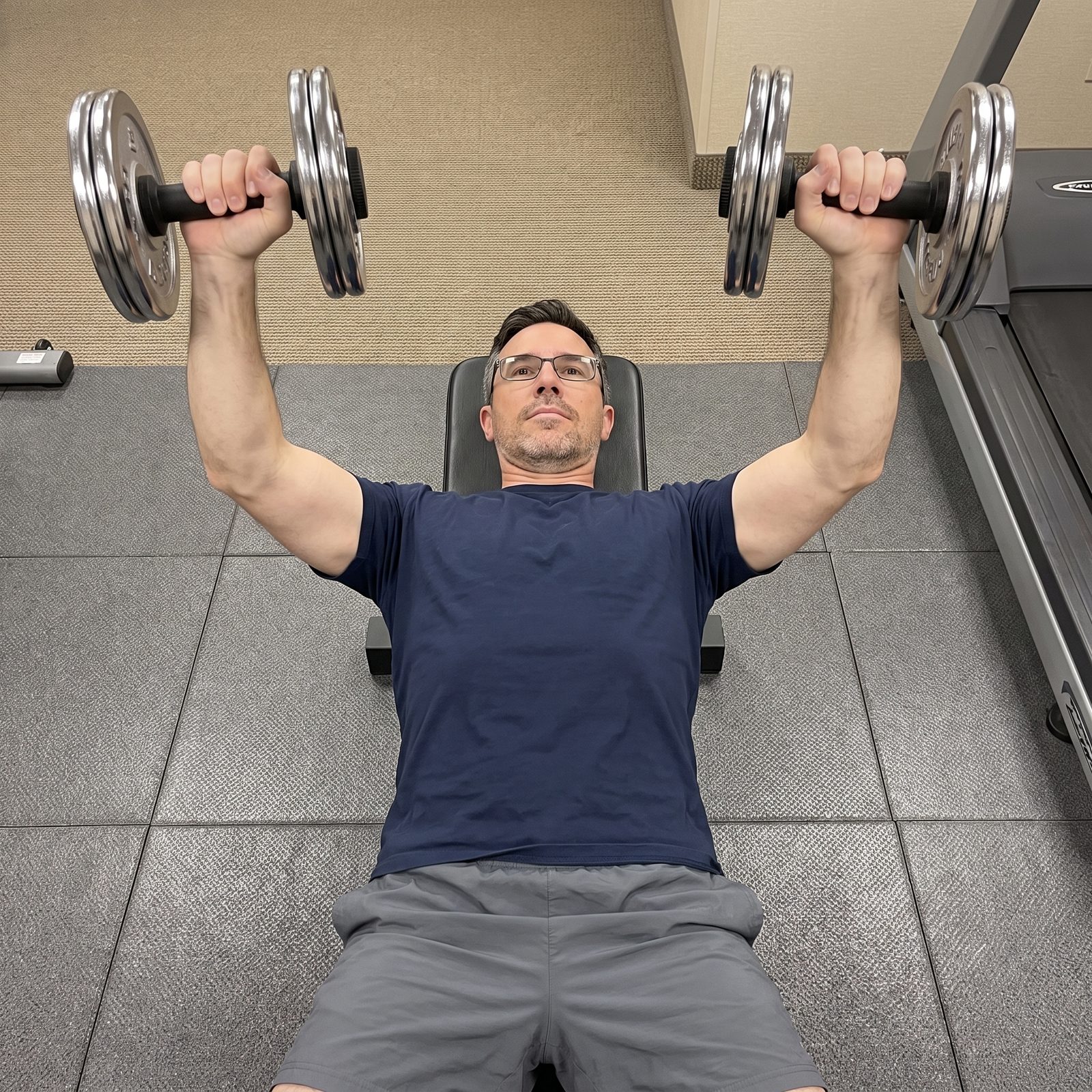 A man in glasses pressing two plate loaded dumbbell handles while lying on a flat bench in a hotel fitness room