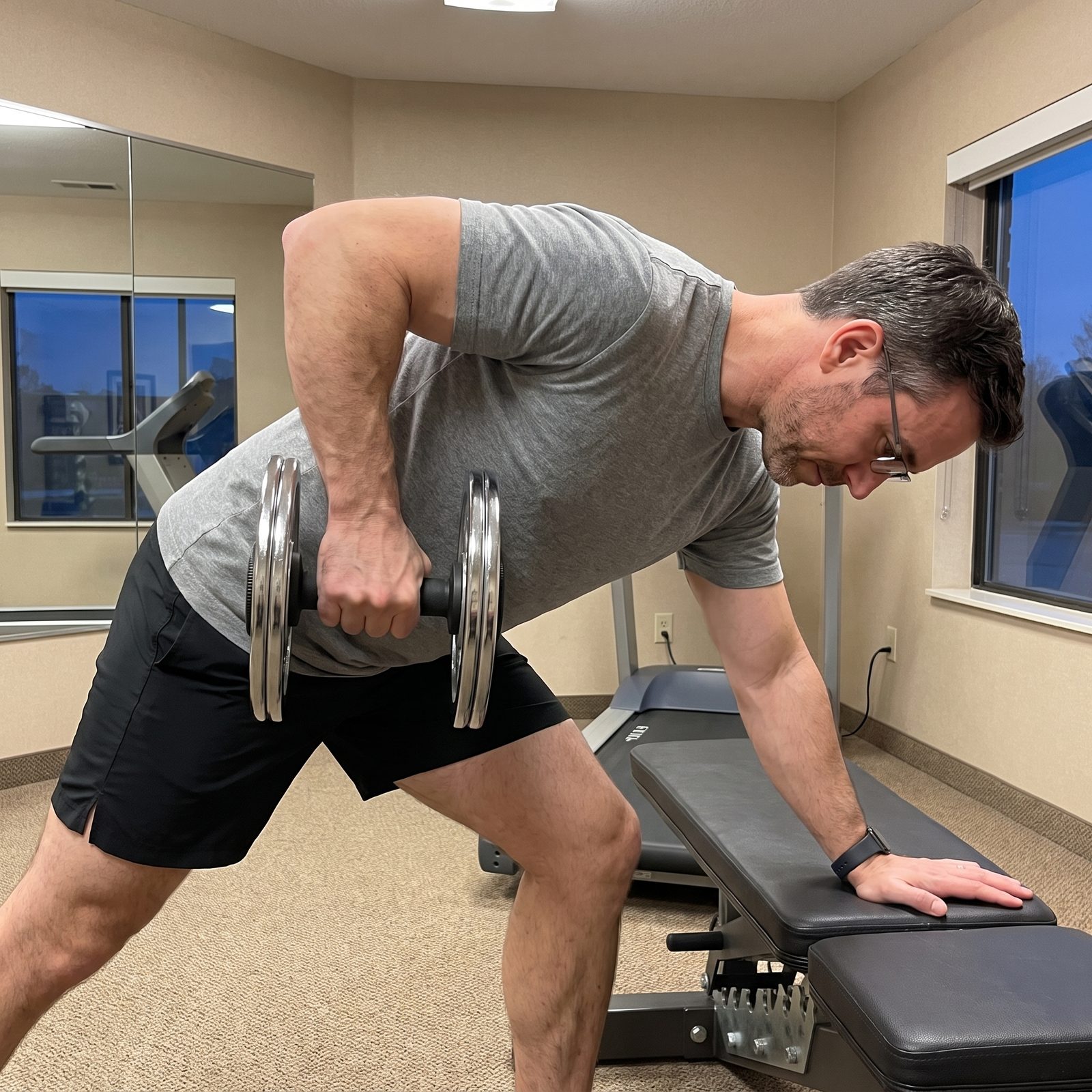 A man in glasses doing a one arm dumbbell row on a hotel gym bench with a plate loaded dumbbell handle carrying two large plates
