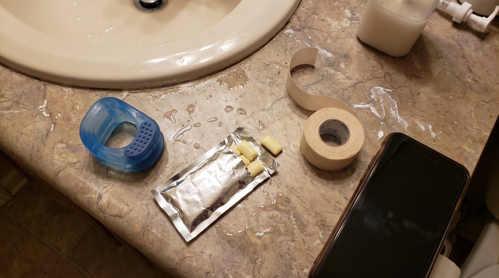 A blue silicone jaw exercise ball, an open foil pack of mastic gum, a roll of mouth tape and a phone face down on a wet bathroom counter