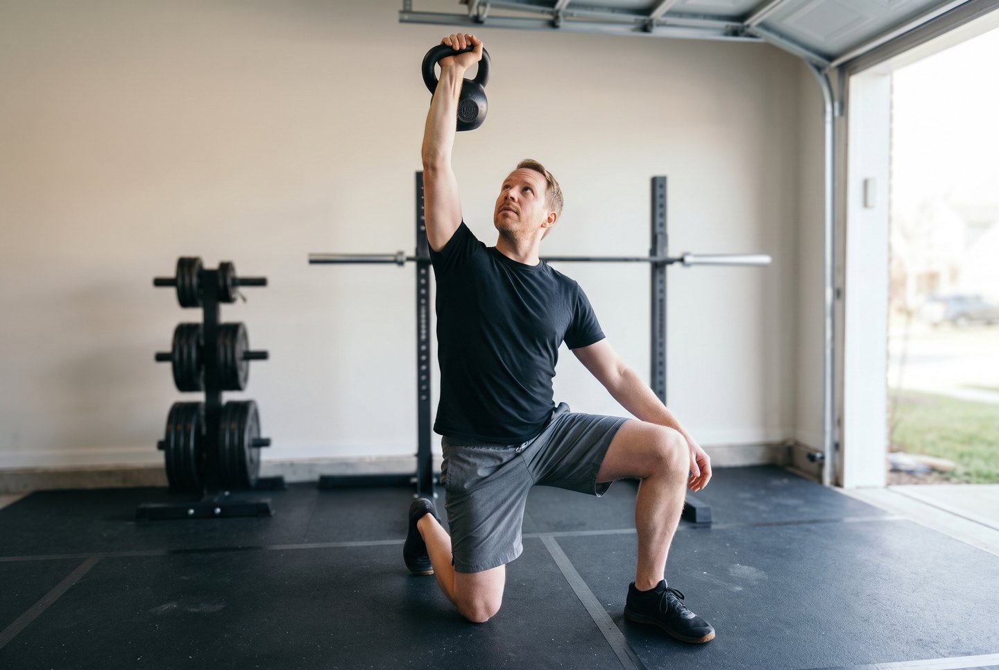A man in the half-kneeling position of a Turkish get-up in a garage gym, a cast-iron kettlebell locked out overhead