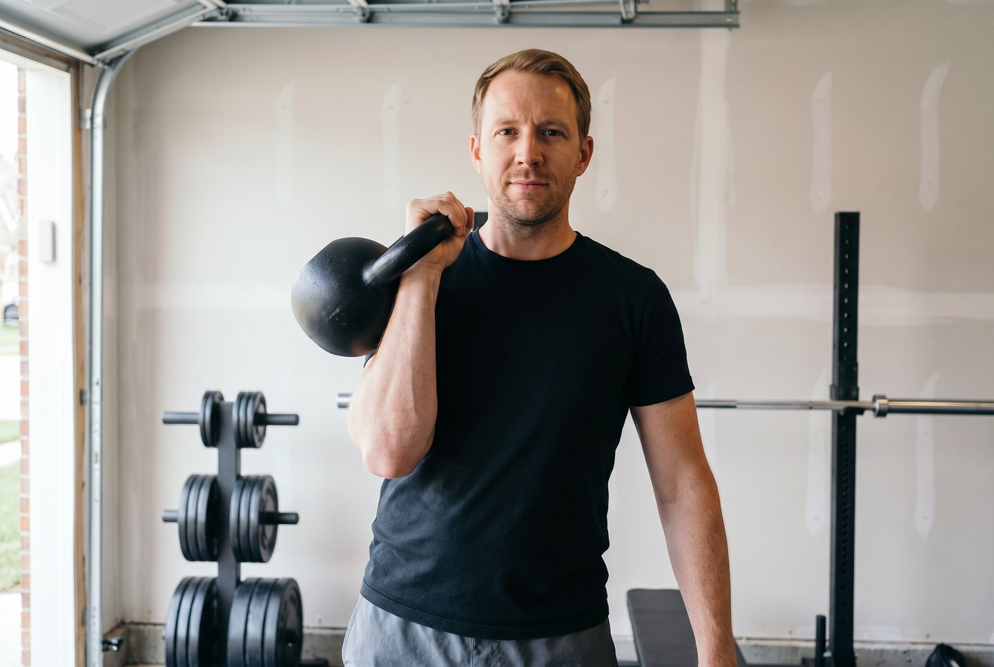 A man in a garage gym with a cast-iron kettlebell cleaned and racked at his shoulder, the bell resting on his forearm