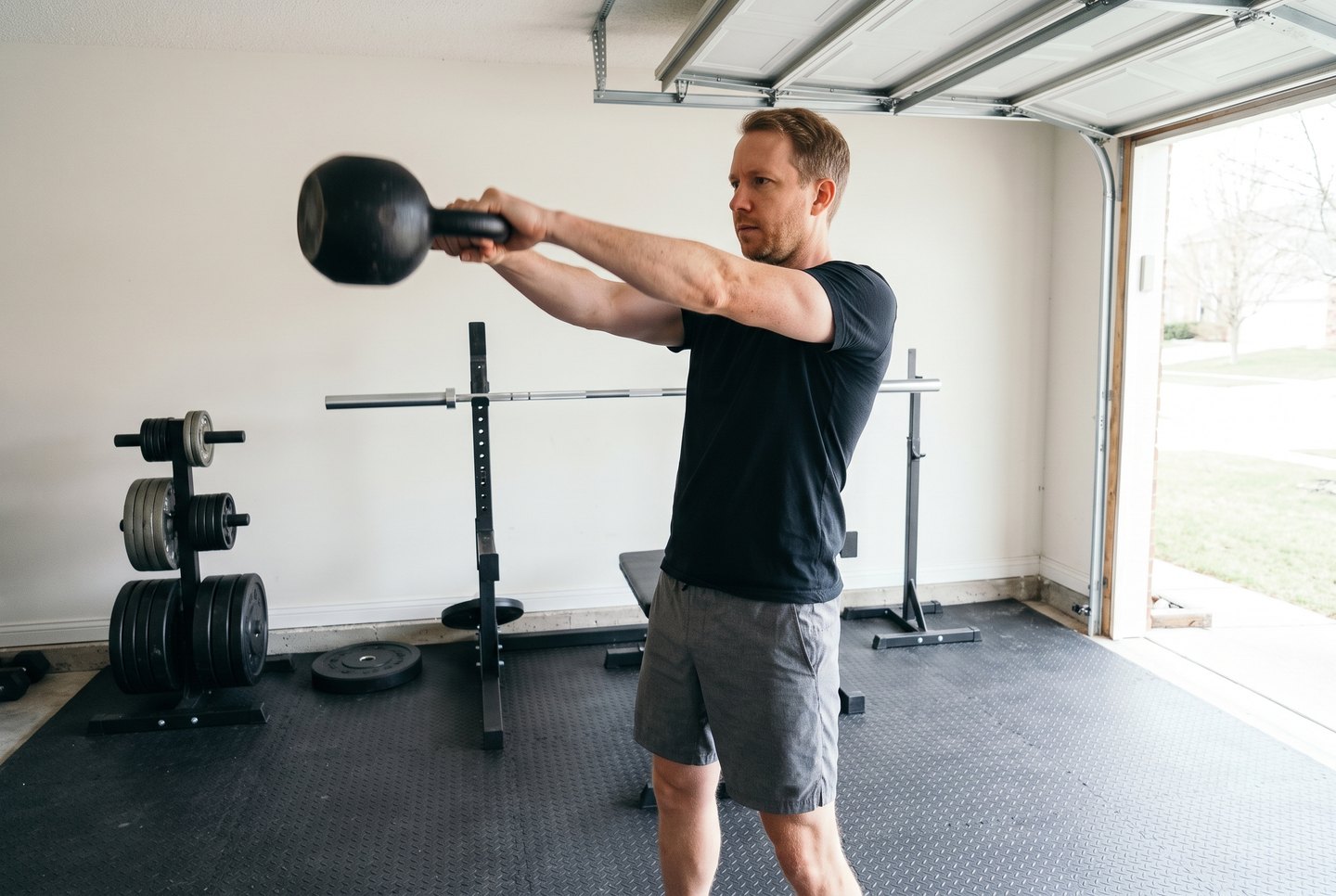 A man warming up with a two-hand kettlebell swing in a garage gym, the garage door open behind him