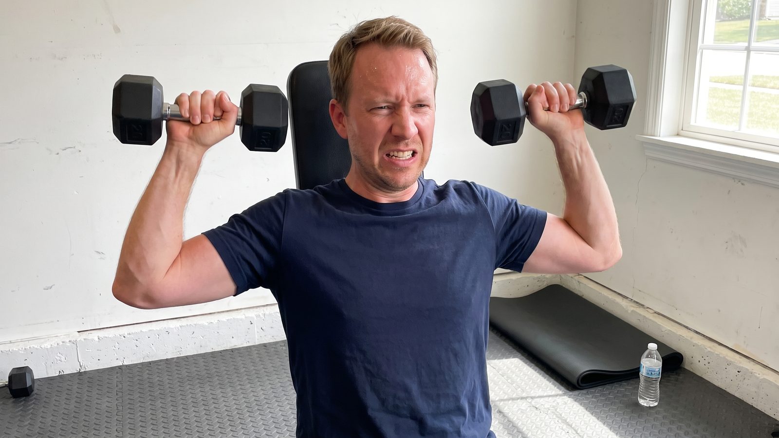 A man in a navy t-shirt seated on an upright bench in a garage gym, pressing two small black hex dumbbells at ear height on the last rep of a set, face grimacing, a rolled mat and water bottle on the floor behind him