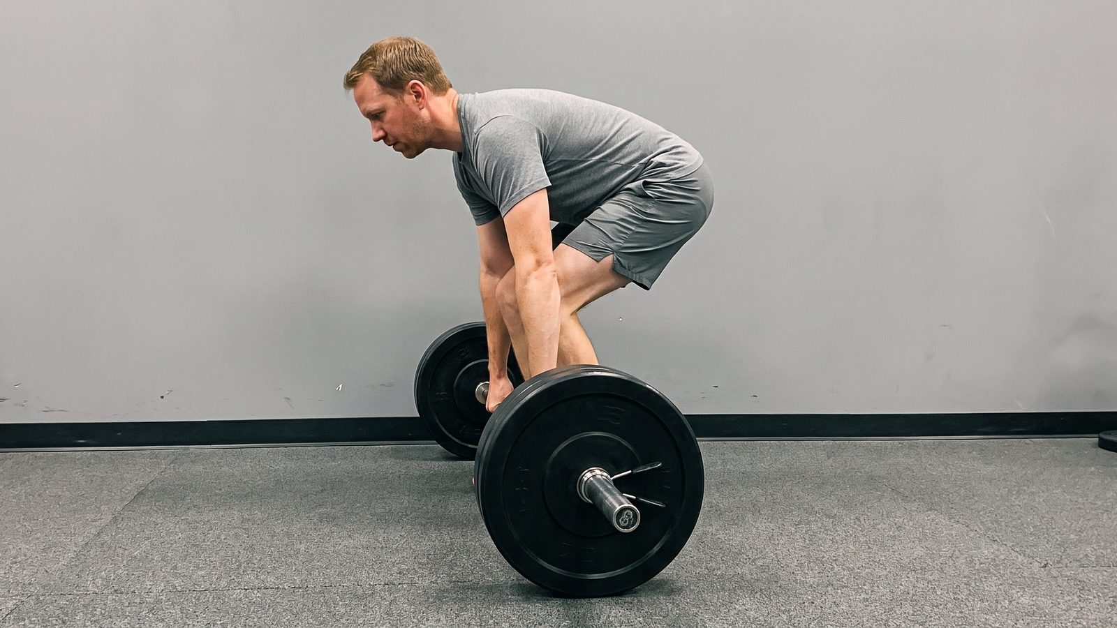 A lifter set up over a loaded barbell for a conventional deadlift, photographed from the side with his arms hanging straight to the bar
