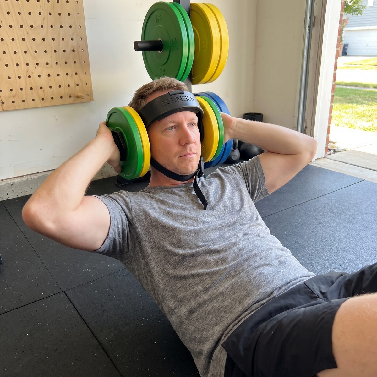 The writer lying on his back on garage floor tiles, lifting his head in a neck curl with the helmet loaded