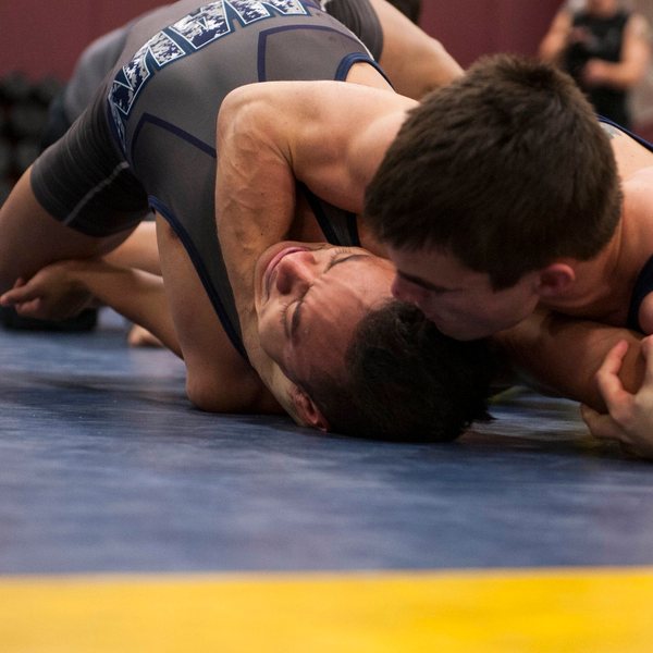 Two wrestlers on a blue mat, the one underneath pinned onto his head and neck by his opponent's weight