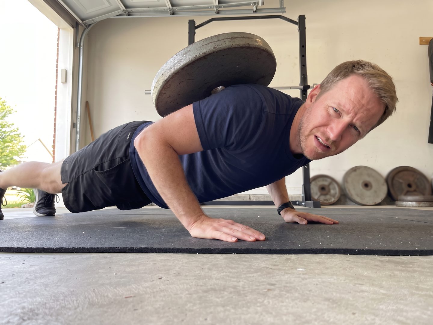 A man at the bottom of a push-up in his garage with an old iron plate tipping off his upper back
