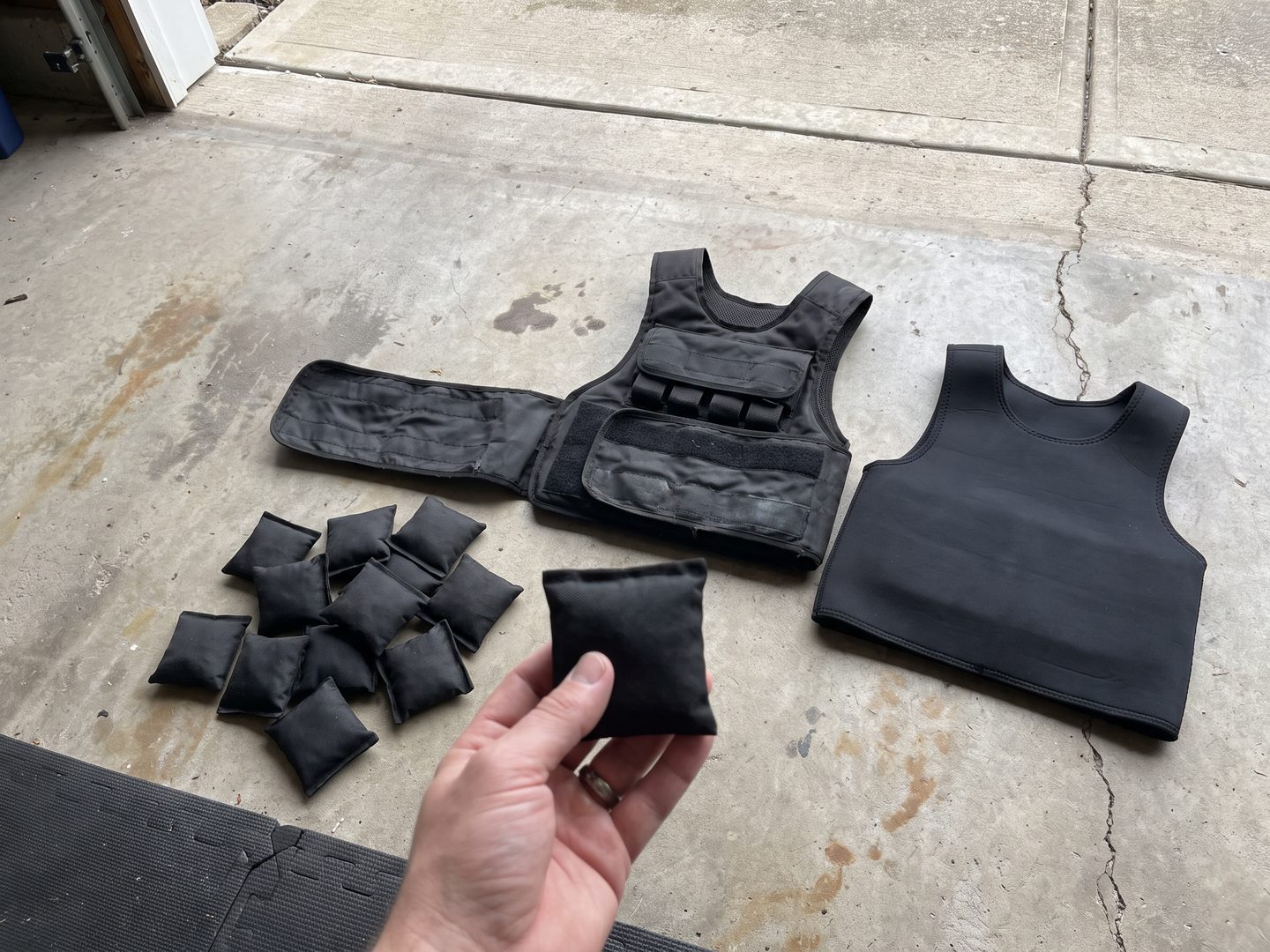 An open adjustable weight vest with a pile of small sand bags spilled beside it and a fixed neoprene vest next to it on a garage floor