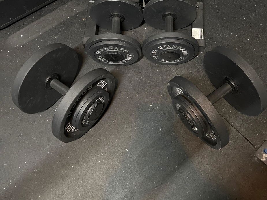 Two pairs of black plate loaded dumbbell handles on a rubber gym floor, loaded with black iron plates