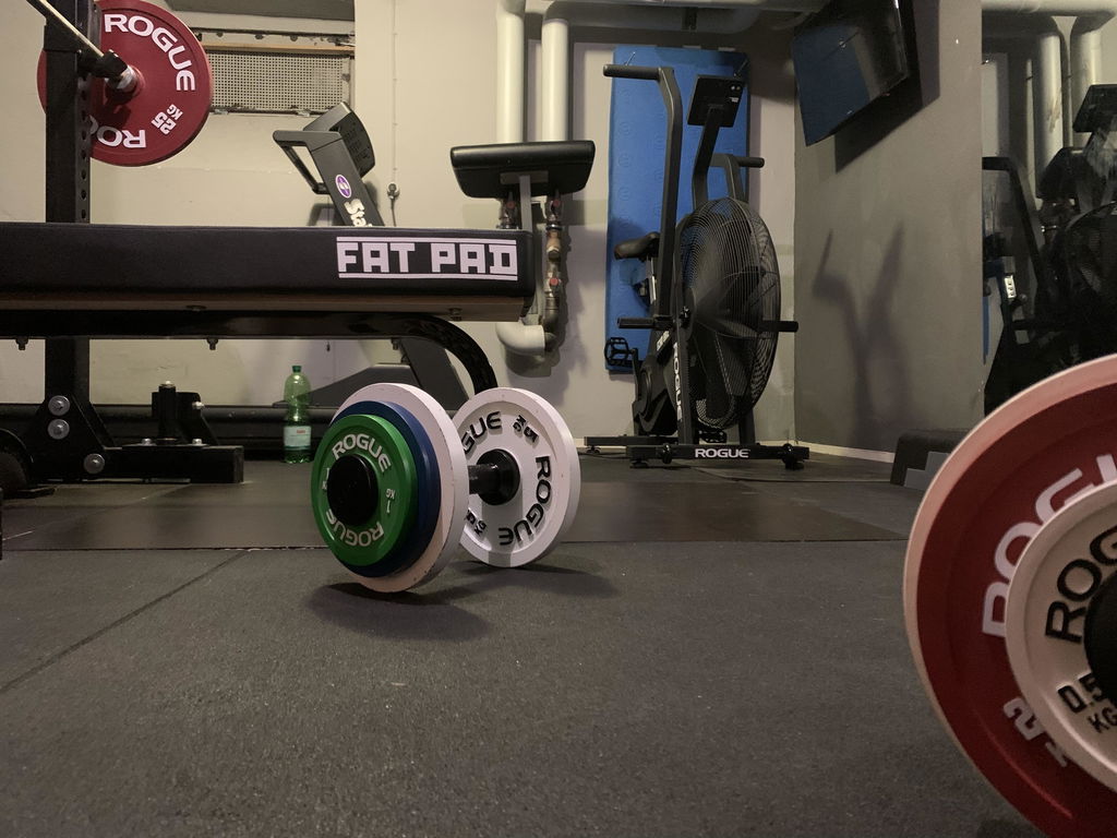 A plate loaded dumbbell handle carrying small rubber change plates on the floor of a commercial gym beside a bench