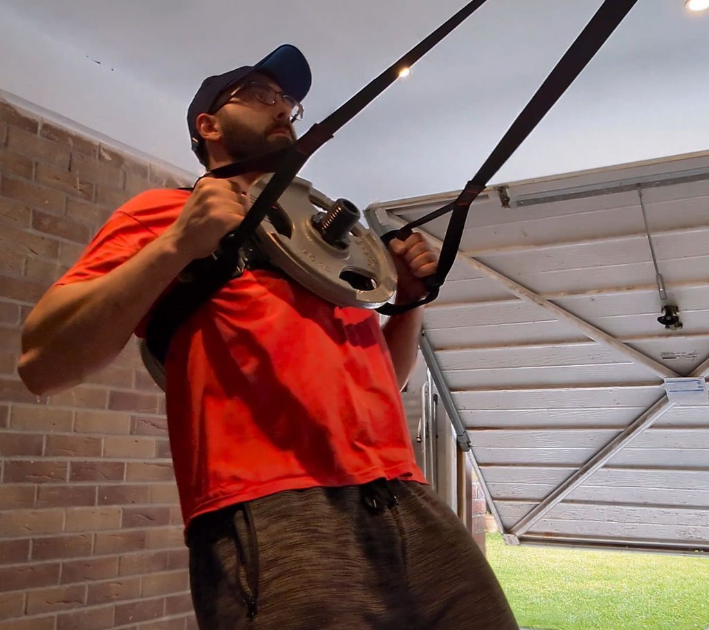 A man in a red shirt doing a pull-up on suspension straps in his backyard wearing a Kensui EZ-VEST loaded with a plate