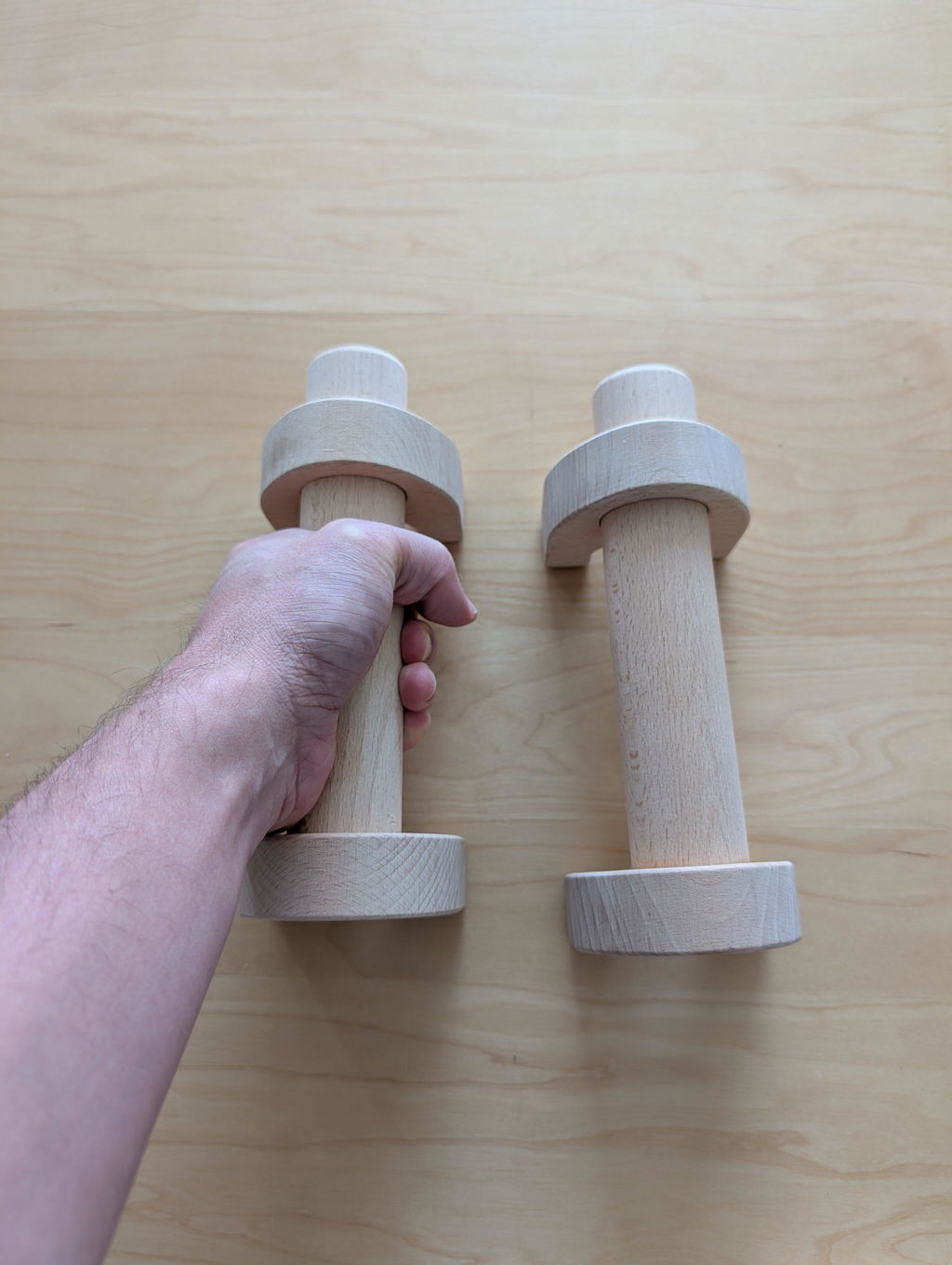 A hand holding one wooden parallette next to its pair on a wooden floor, showing the size