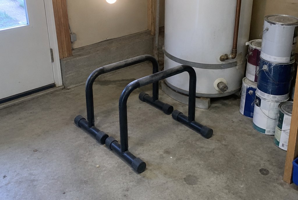 A pair of heavy black steel parallettes standing dusty in a garage corner next to a water heater