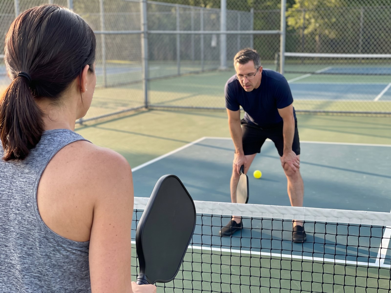The author bent over with a hand on his knee across the net from his wife, who stands in the foreground with her back to the camera, on a public pickleball court in late afternoon sun