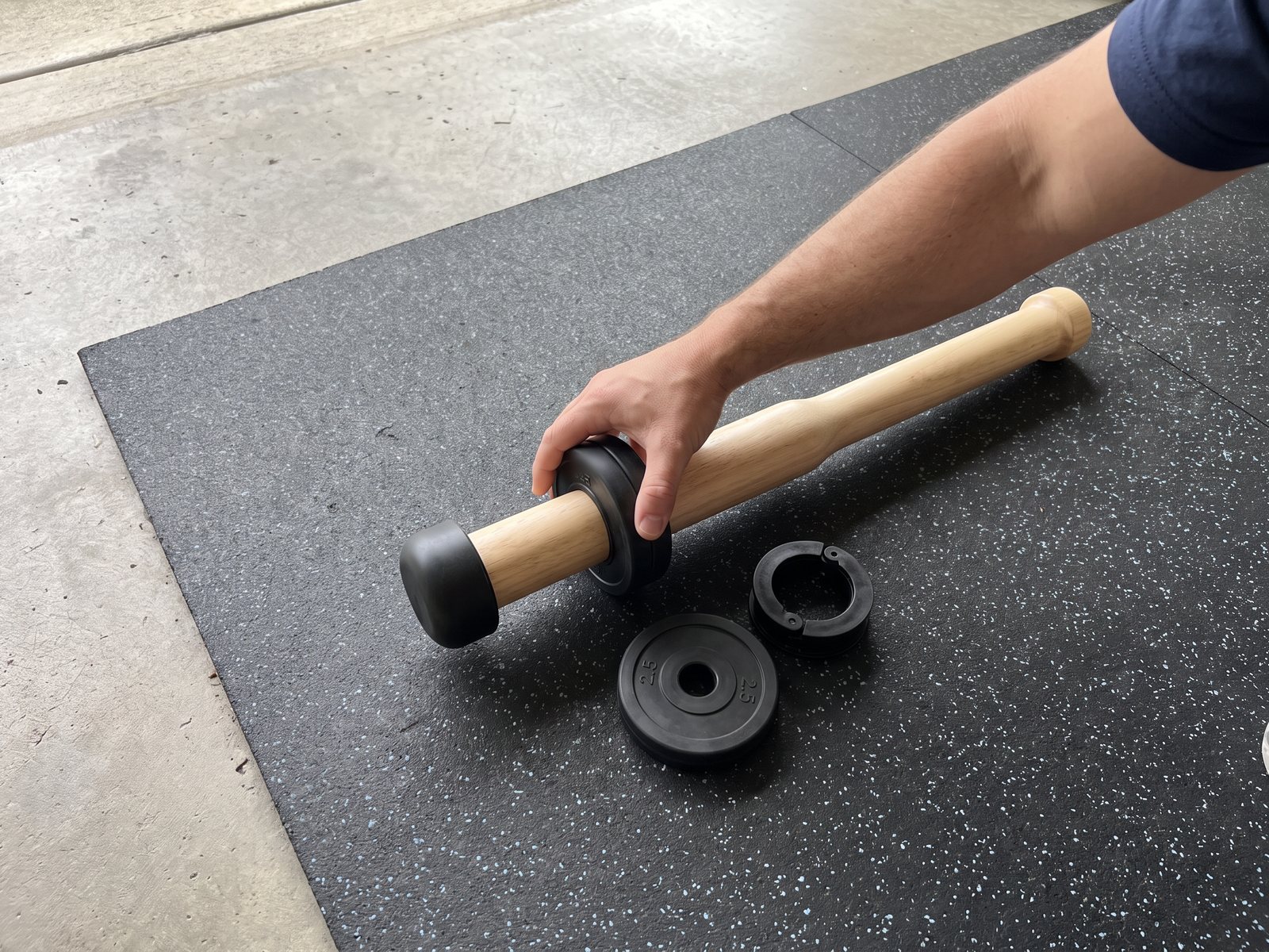 A hand sliding a small black weight plate onto the wooden club's sleeve on a garage mat, a second plate and the open collar beside it