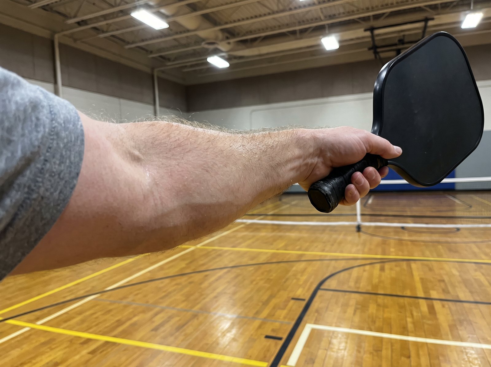 A forearm holding a pickleball paddle straight out at arm's length on an indoor court, wrist cocked, tendons showing