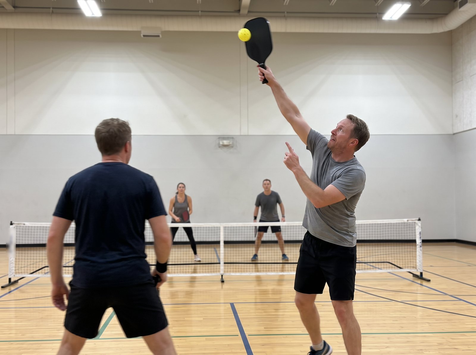 The author reaching up for an overhead on an indoor pickleball court late at night, two opponents across the net