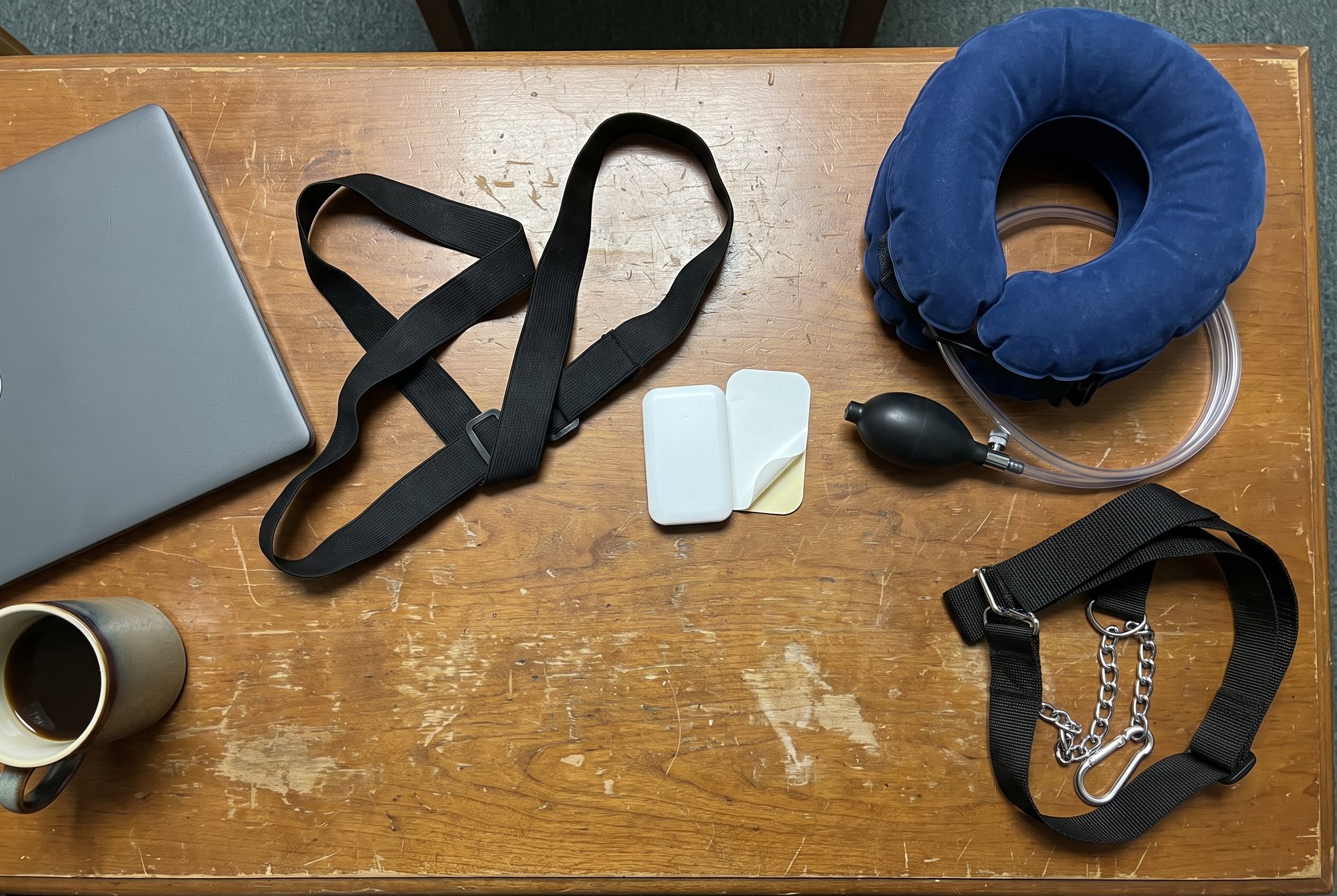 Laid out on a wooden desk beside a laptop: a black figure-eight posture strap, a small white stick-on posture sensor, a blue inflatable neck traction collar with a hand pump, and a black nylon head strap with a steel chain