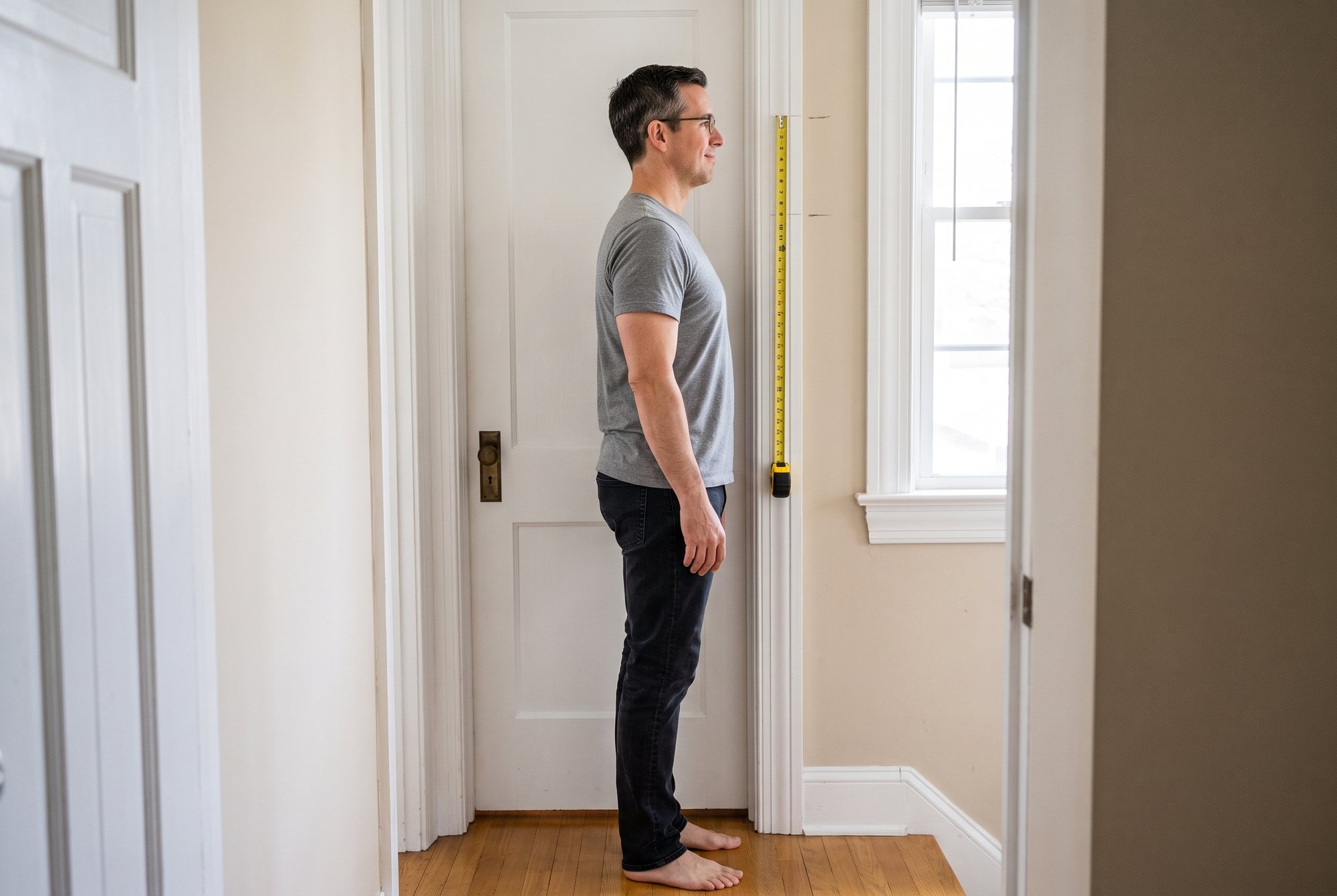 A man in a grey t-shirt standing tall and relaxed against a white door frame, head level over his shoulders, a yellow tape measure hanging beside two pencil marks level with the top of his head