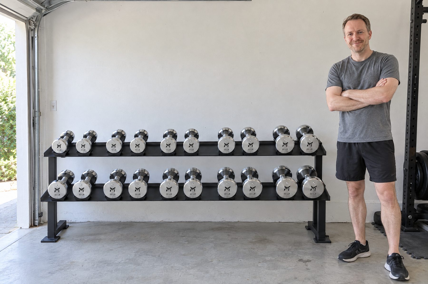 A man standing with his arms folded beside a two tier black rack holding ten pairs of chrome dumbbells in a garage gym