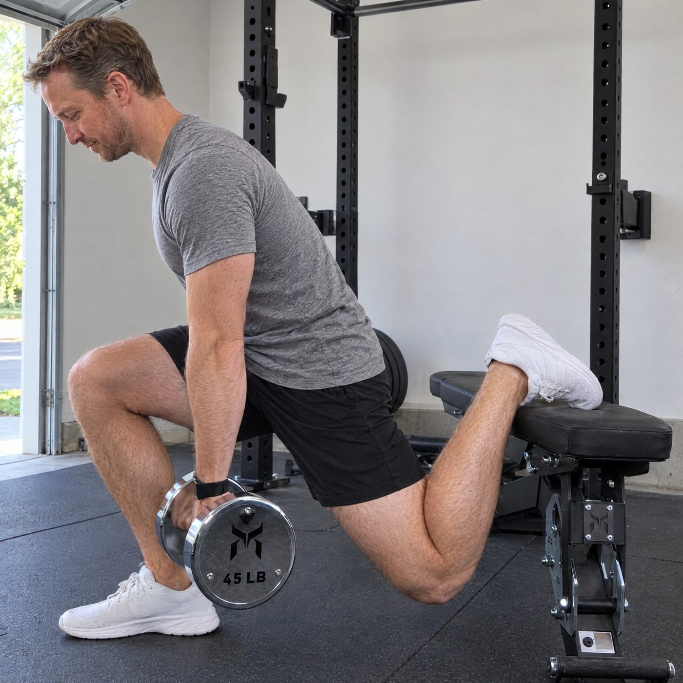 A man doing a rear foot elevated split squat in a garage gym holding a chrome 45 pound dumbbell at his side