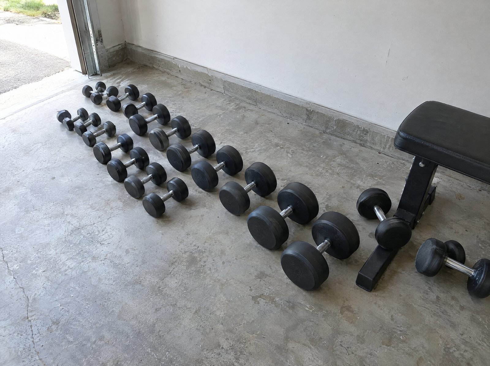 Two crooked lines of black round dumbbells left on a concrete garage floor with no rack, a few rolled against a bench