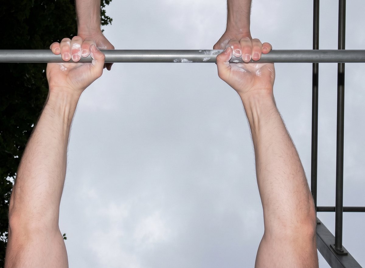 Looking straight up at two hands gripping a plain steel bar with palms facing forward, against a grey sky