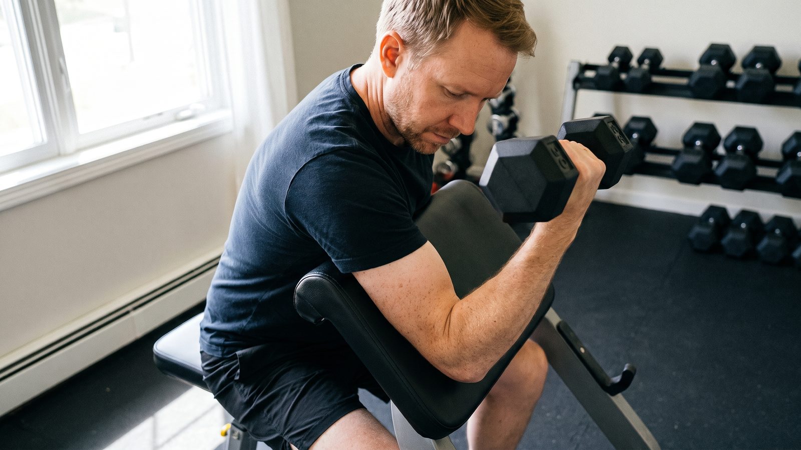 A man in a navy t-shirt seated at a preacher curl bench in a home gym, upper arm on the angled pad, curling a black hex dumbbell and looking down at his own biceps, a rack of dumbbells behind him