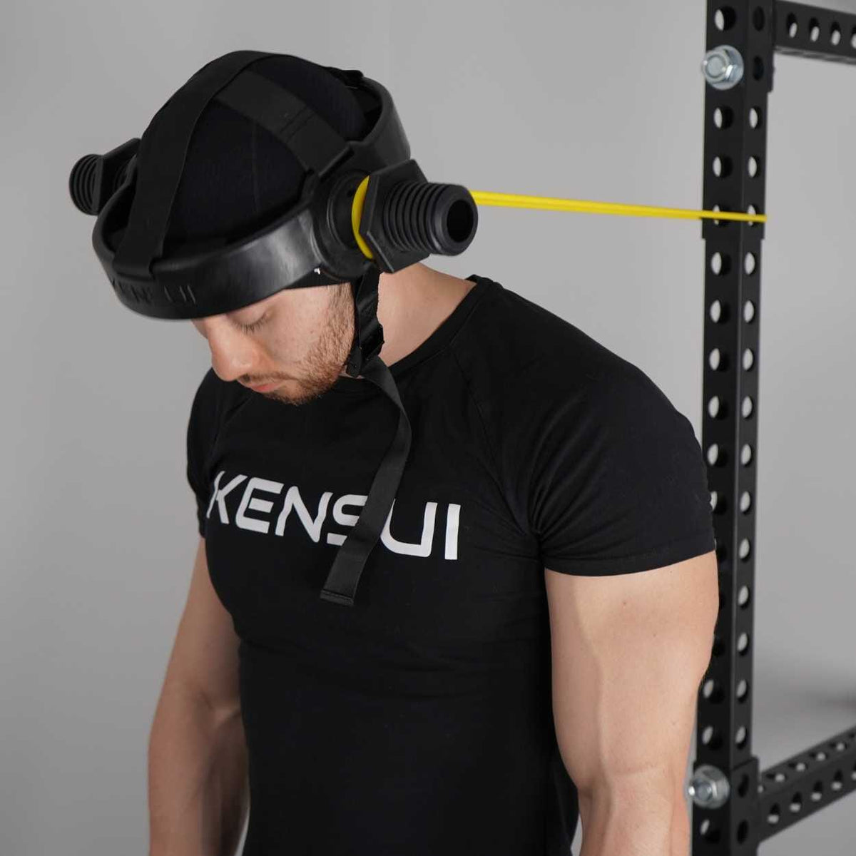 A man in a black t-shirt wearing the neck helmet with the chin strap fastened, a yellow band clipped to the ear sleeve, doing a slow neck curl in a squat rack