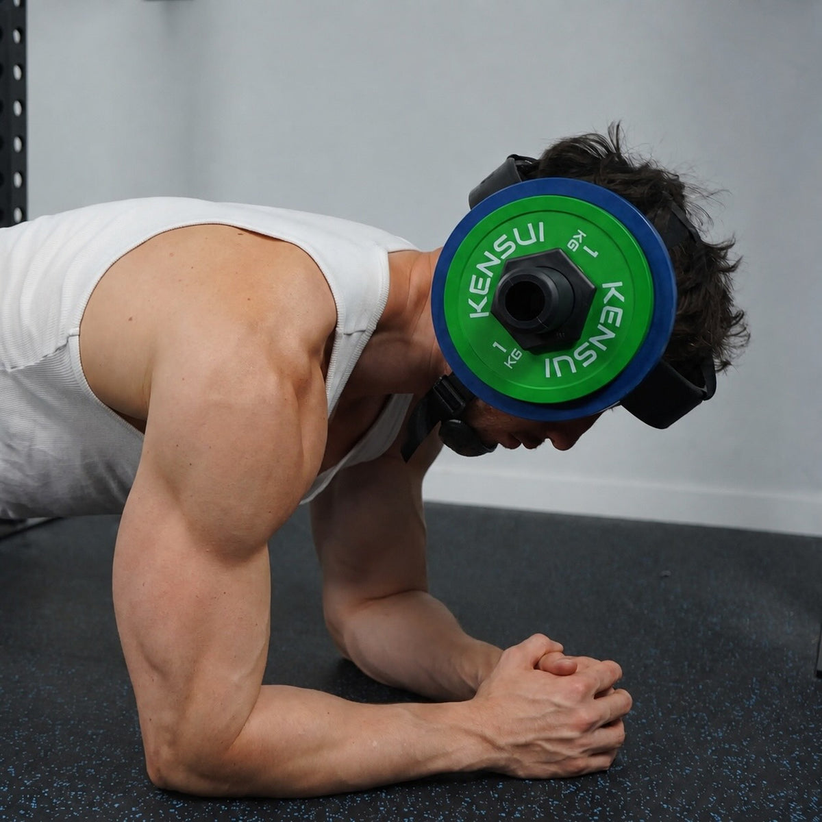 A man in a white vest holding a plank on a gym floor with the neck helmet on, a green plate at each ear, head held level against the load