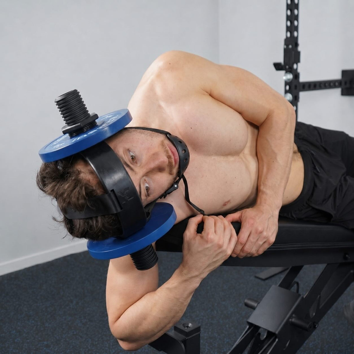 A man lying on his side on a bench doing a slow side bend with the loaded neck helmet on, a blue plate at each ear